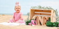 a baby girl in a pink tiara sits on the sand next to a wooden crate